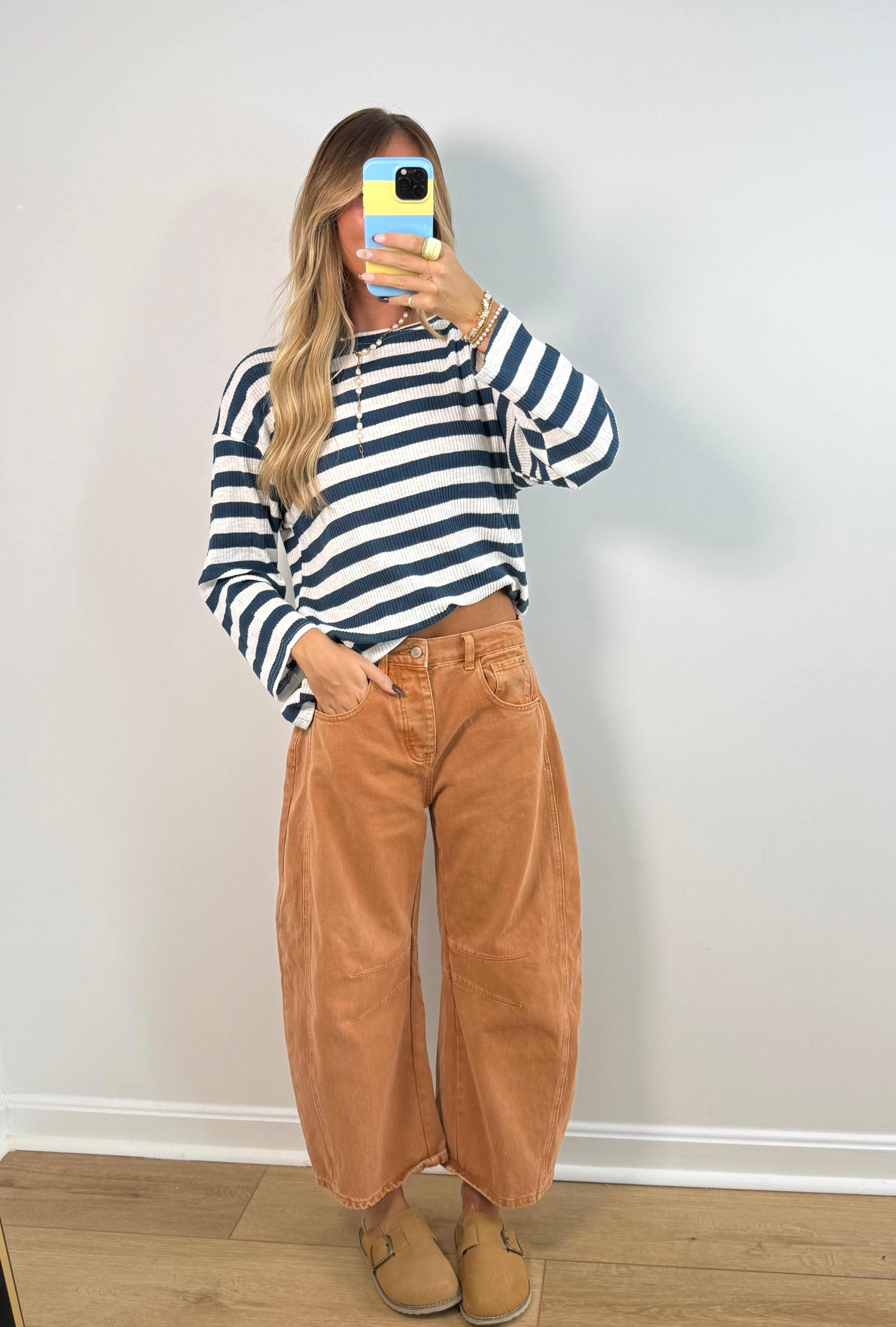 A woman takes a mirror selfie wearing a navy and white striped long-sleeve henley shirt, high-waisted wide-leg tan corduroy pants, and tan slip-on shoes against a plain white wall.
