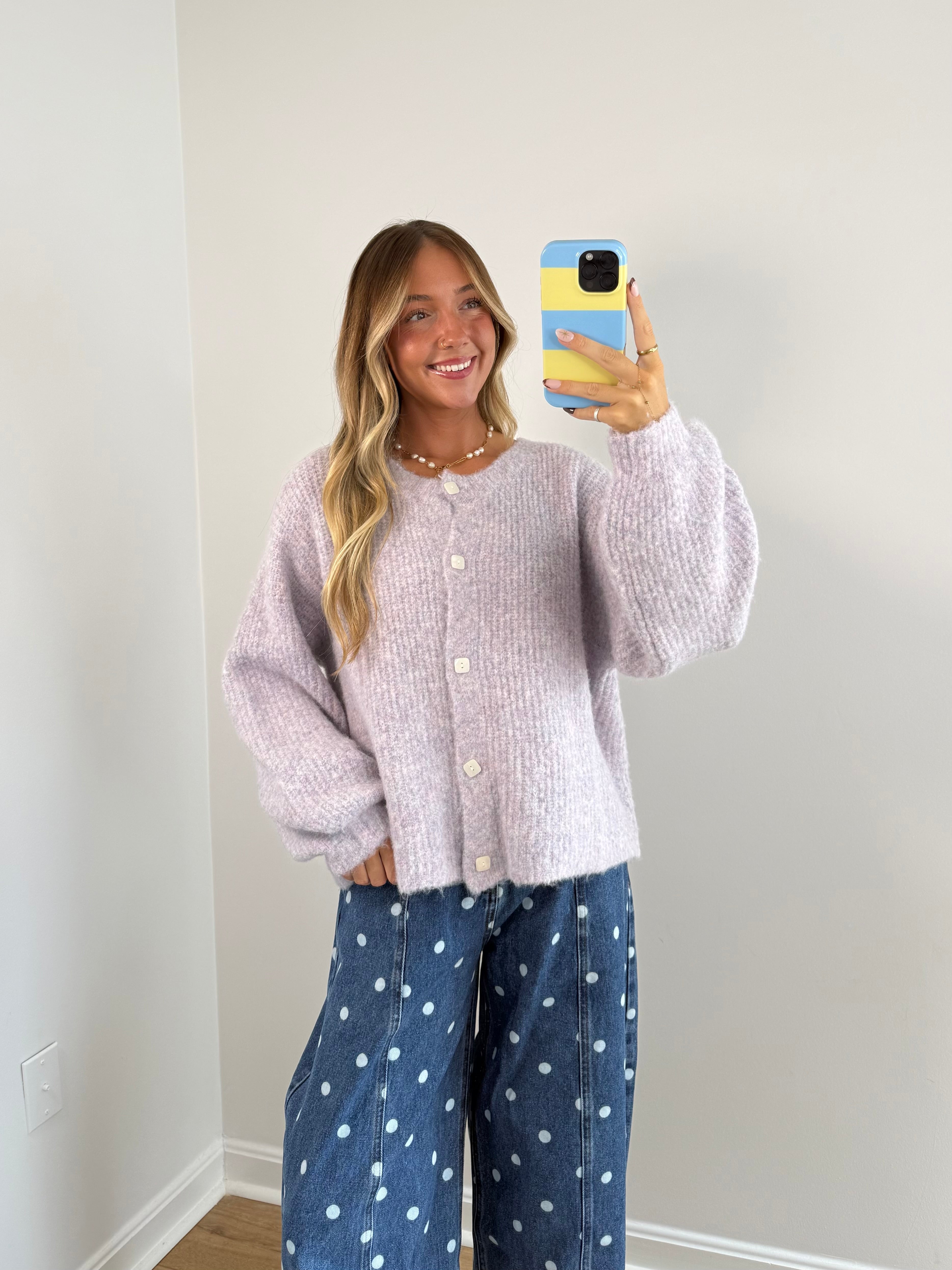 A woman takes a mirror selfie wearing a light lavender fuzzy knit cardigan and blue wide-leg jeans with white polka dots, standing against a plain off-white wall.