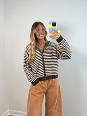 A woman taking a mirror selfie wearing a brown and cream striped zip-up knit sweater and tan corduroy pants, holding a blue and yellow phone case against a plain light gray wall.